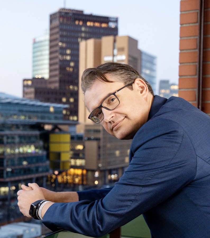 Porträt eines Mannes mit Brille auf einem Balkon vor der Berliner Skyline
